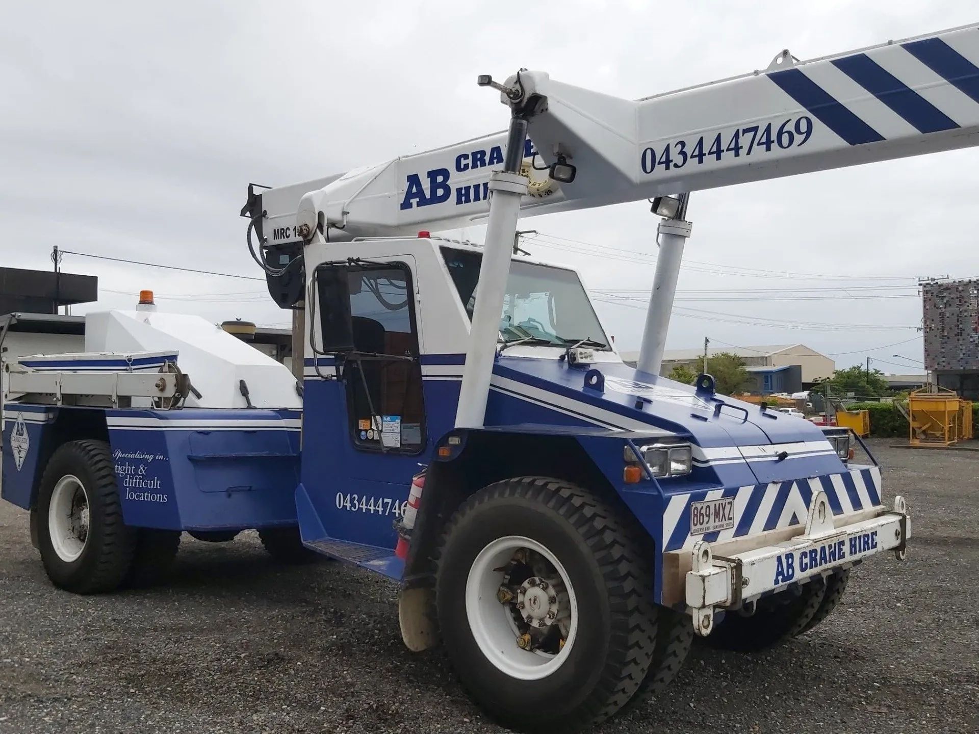 Blue and white tow truck with crane arm parked in a gravel lot under cloudy sky.