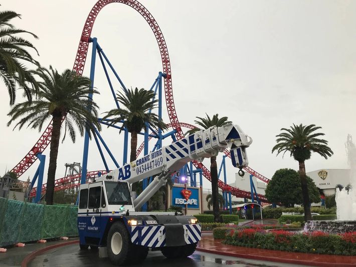 Blue-and-white amusement park vehicle beneath a red roller coaster, with palm trees and flower beds.