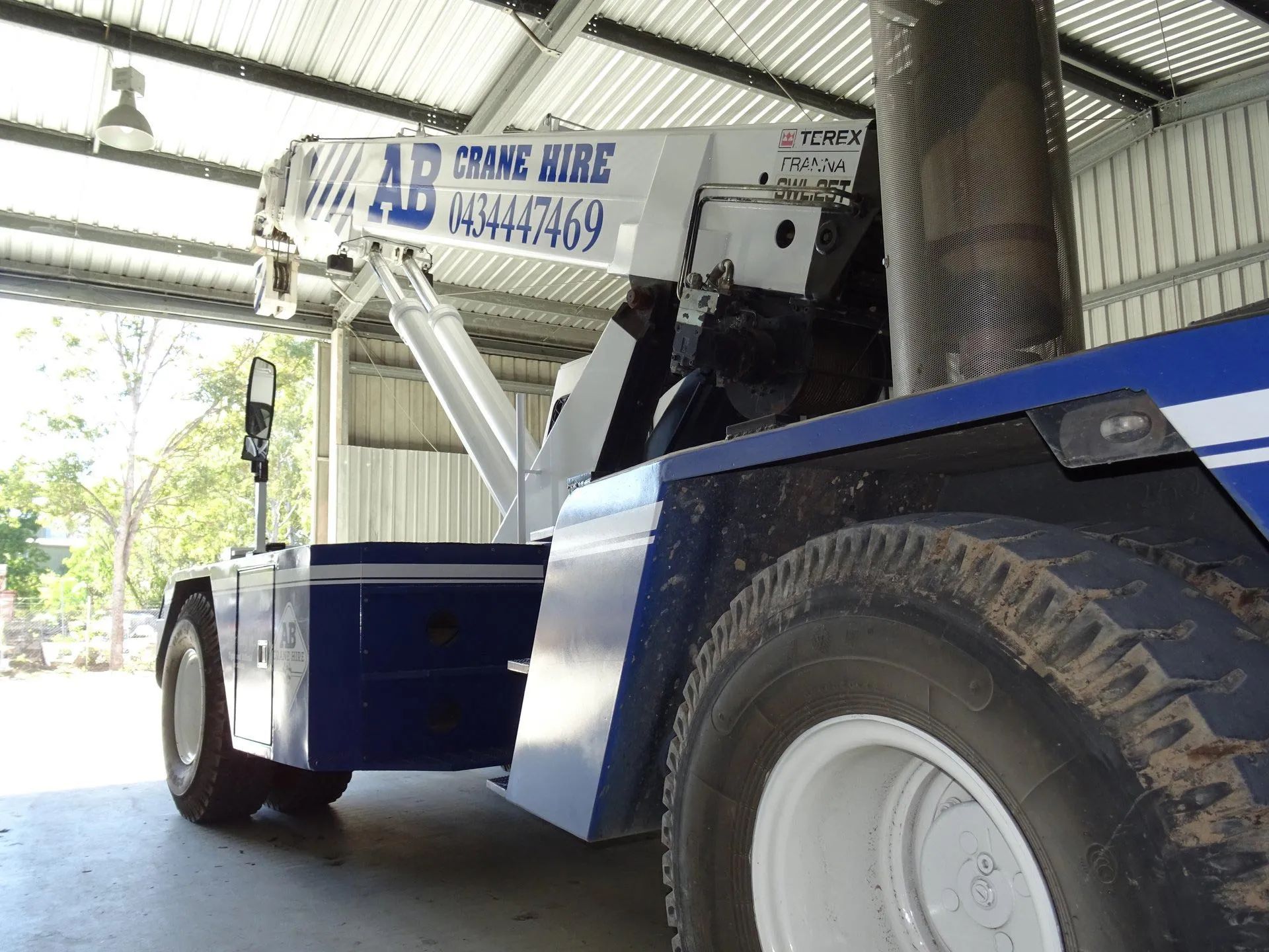 Blue industrial vehicle with large tires parked under a metal-roofed shelter, showing a front-mounted boom arm.
