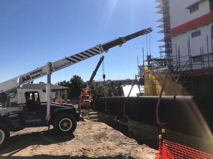 Crane truck lifting a large black pipe at a construction site beside a building.