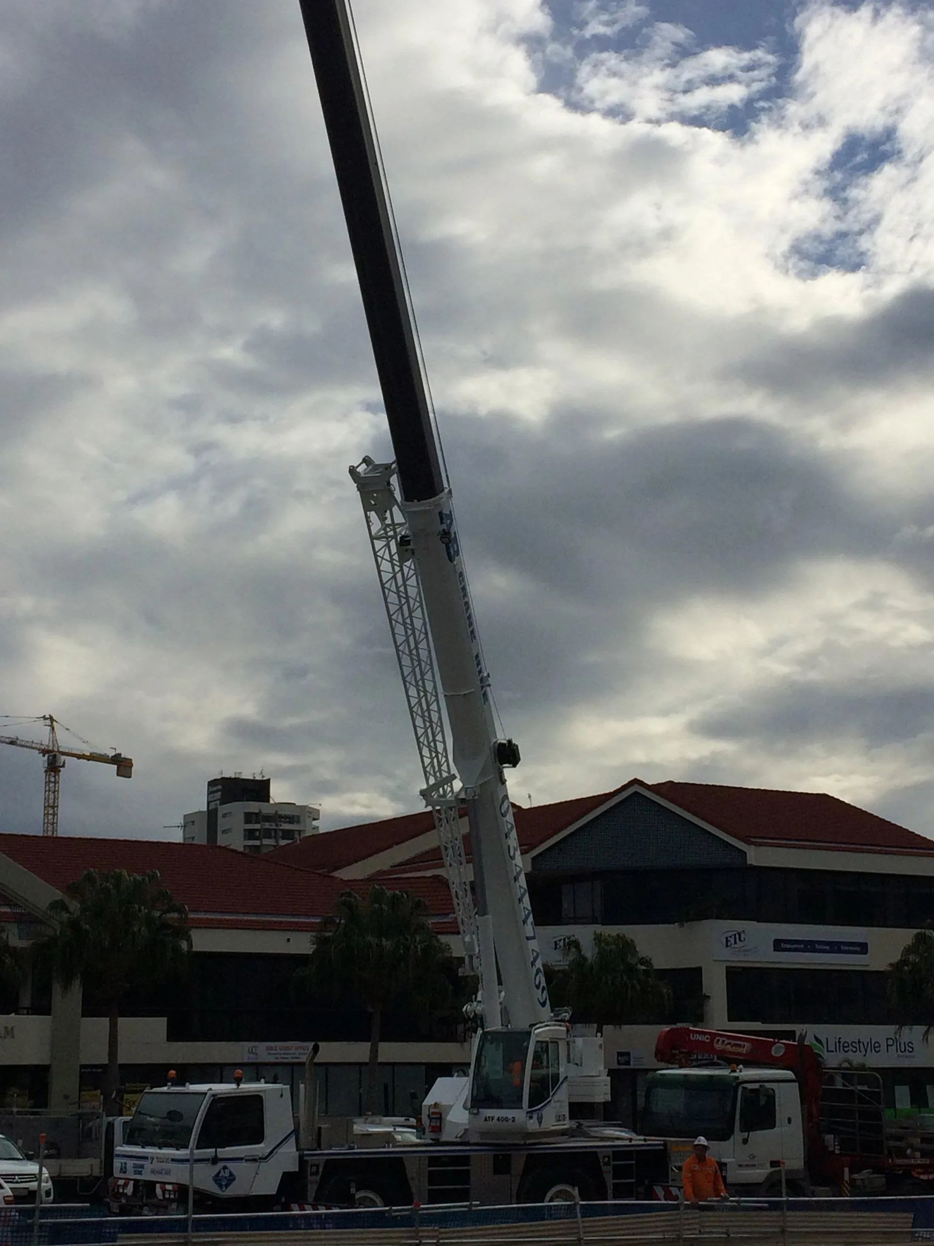 Crane truck lifting a long boom beside a red-roofed building under cloudy skies