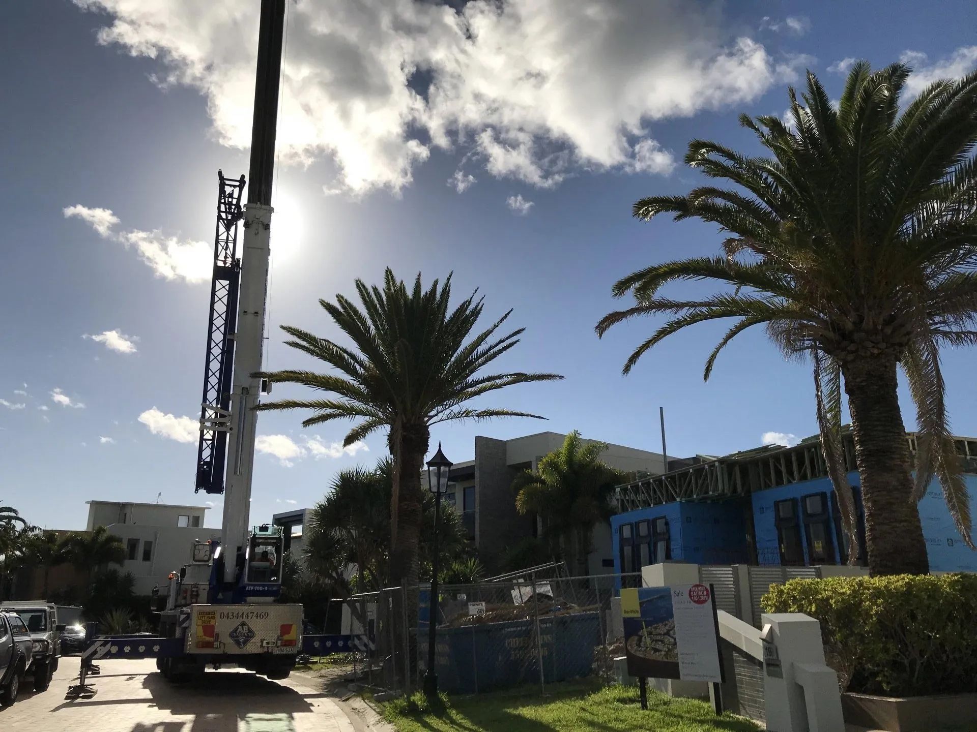 Crane truck on a sunny street beside palm trees and a fenced construction site