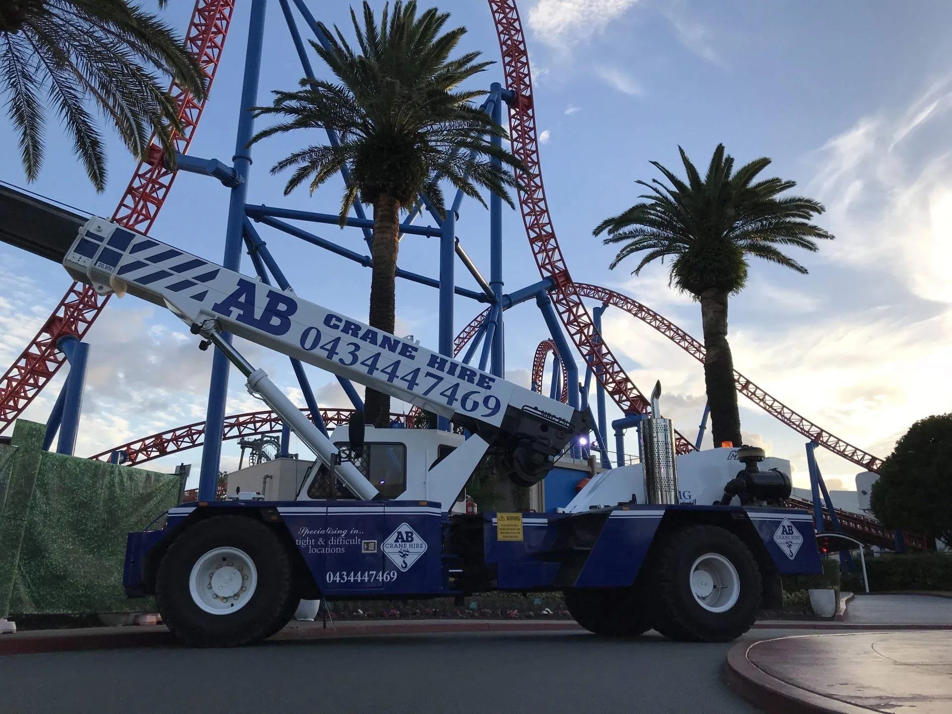 Blue crane truck lifting a large carnival ride frame with palm trees in the background