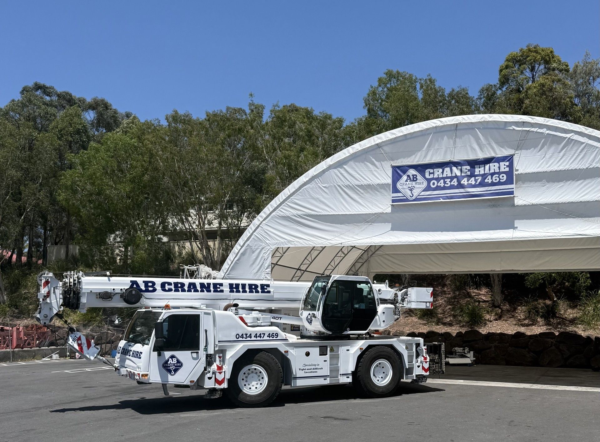 White crane truck parked by a tented building with a banner, under trees on a sunny day