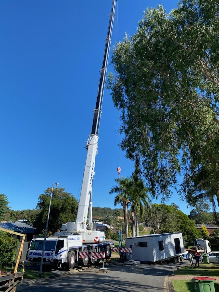 Crane lifts a long black-and-white mast in a sunny yard with trucks, a trailer, and trees.