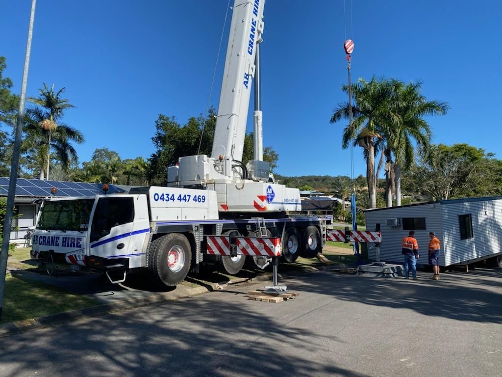 Large white crane truck on a roadside with workers, palm trees, and a small building nearby