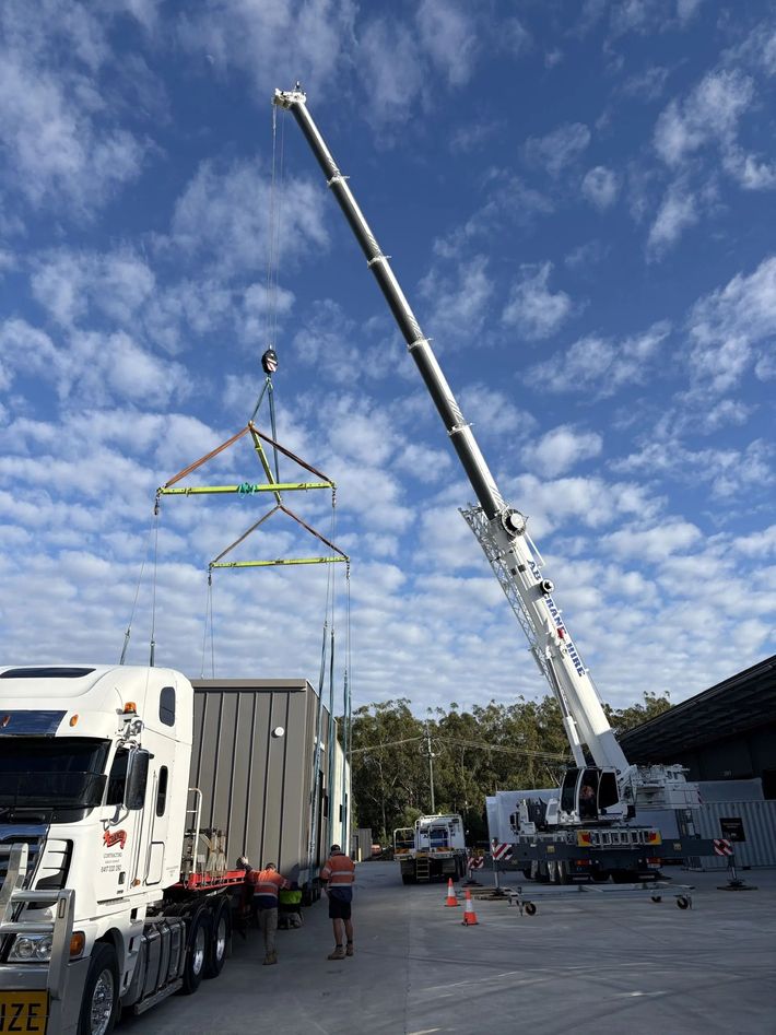 Crane lifting a steel truss beside trucks and workers at a construction site under a blue sky