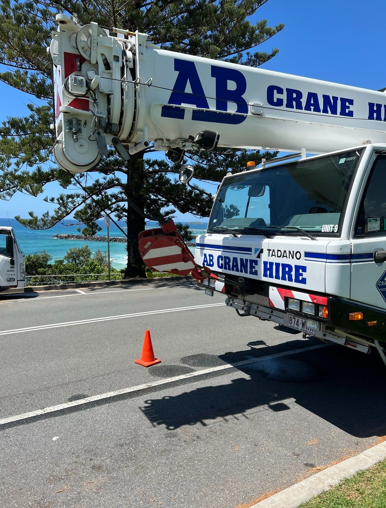 Crane truck parked by the roadside with an orange cone beside it, overlooking a blue ocean.