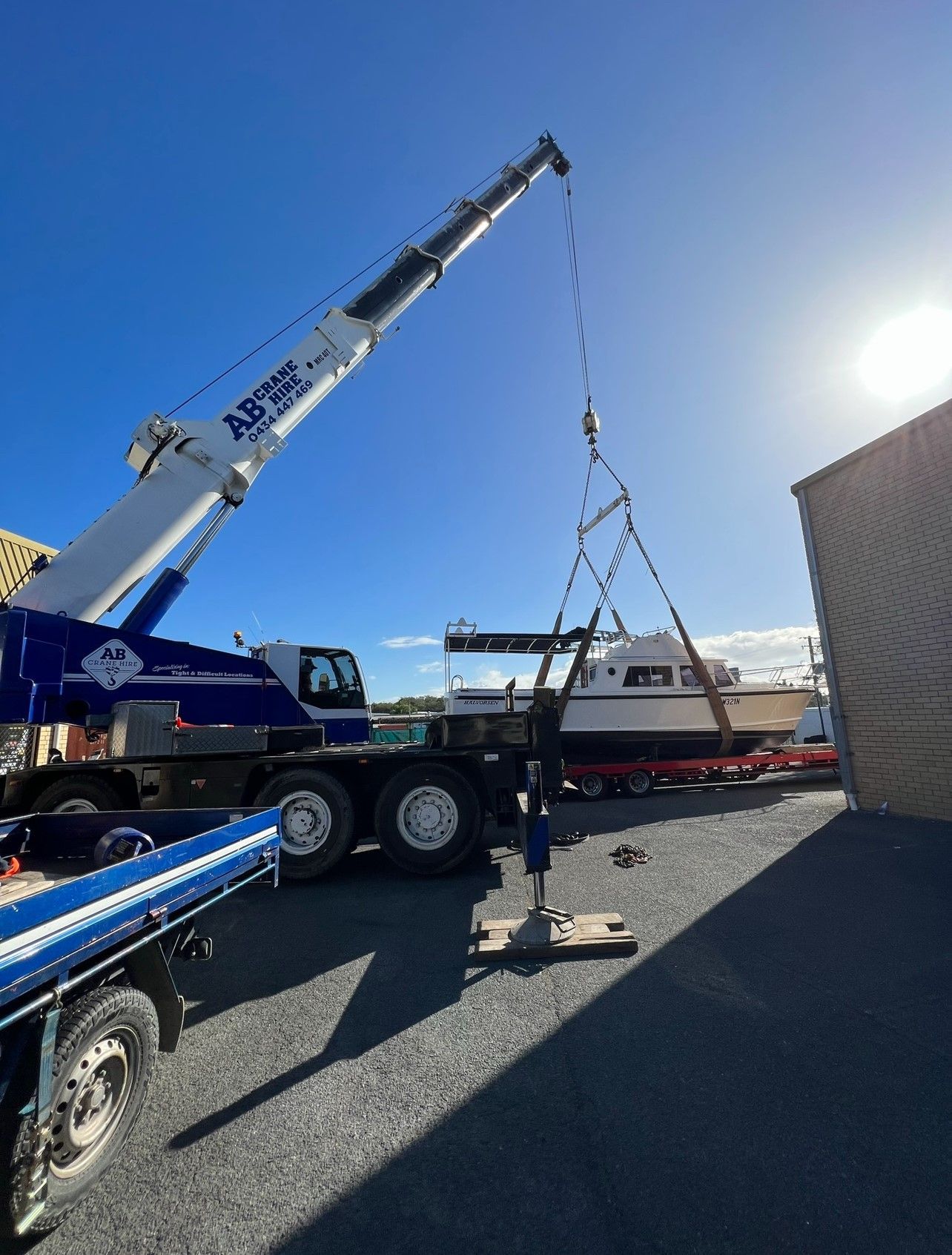 Crane lifting a red boat trailer in a sunny gravel lot beside a building.