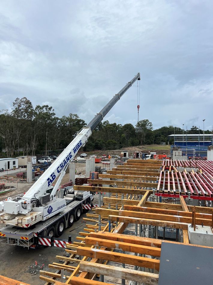 Large crane at a construction site lifting a beam beside rows of wooden framing and concrete forms