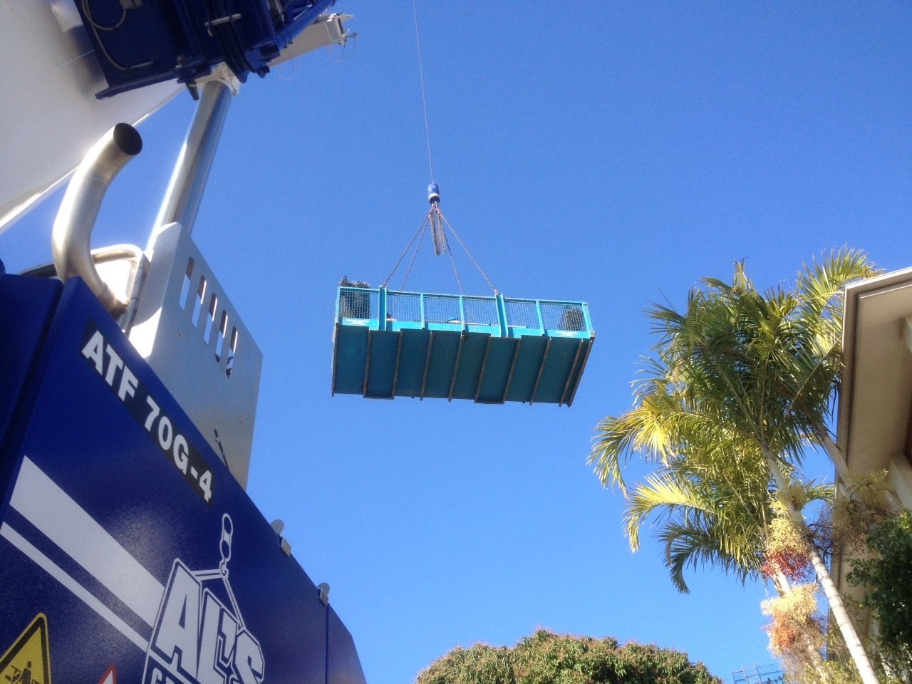 Blue cargo load suspended from a crane beside a building and palm tree against a clear sky.