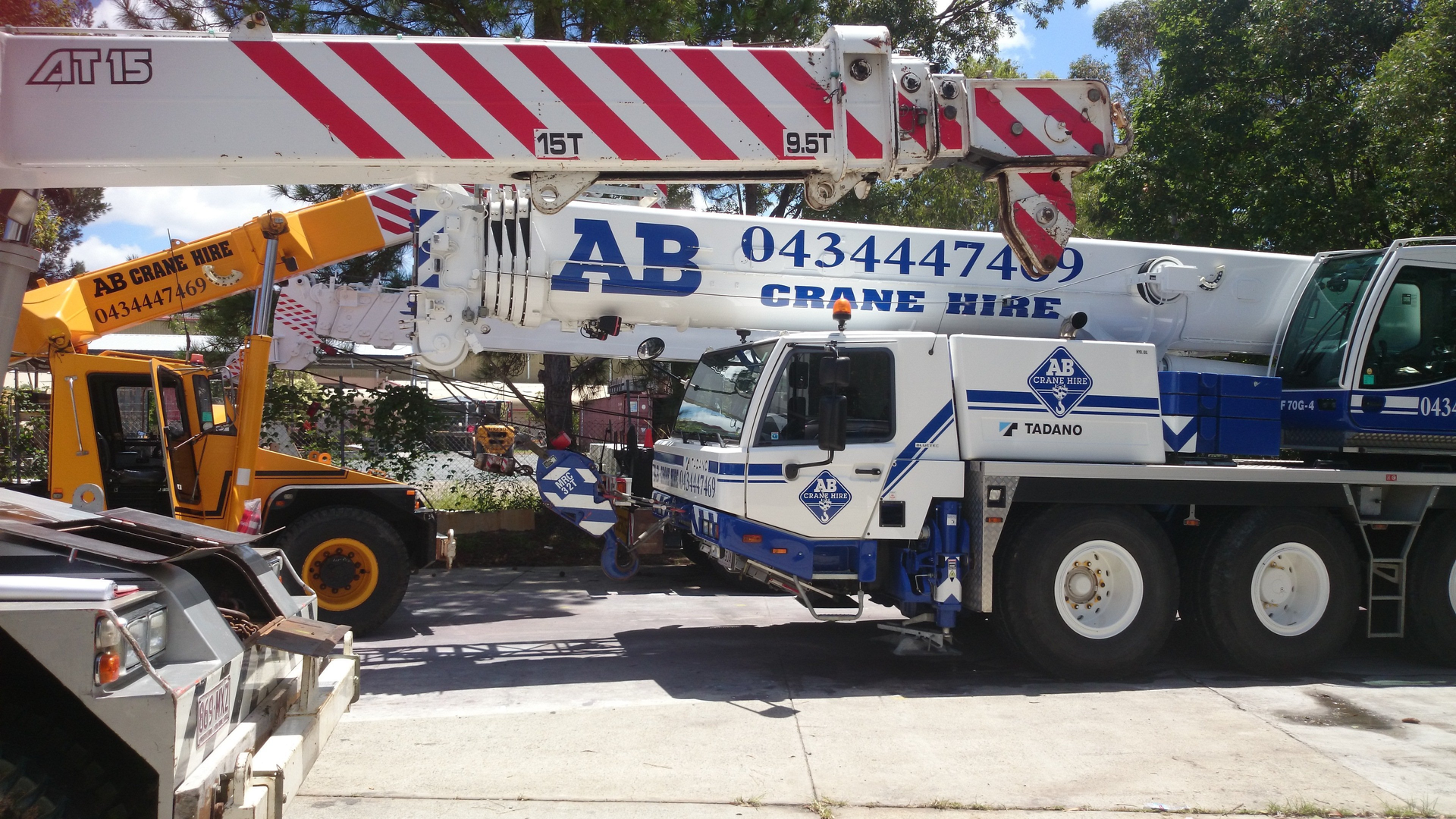 Two cranes lifting a striped hazard-marked beam on a street, with a blue crane truck in front