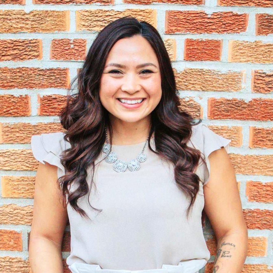 Smiling woman in a light gray top, standing before a brick wall with a necklace and wavy dark hair