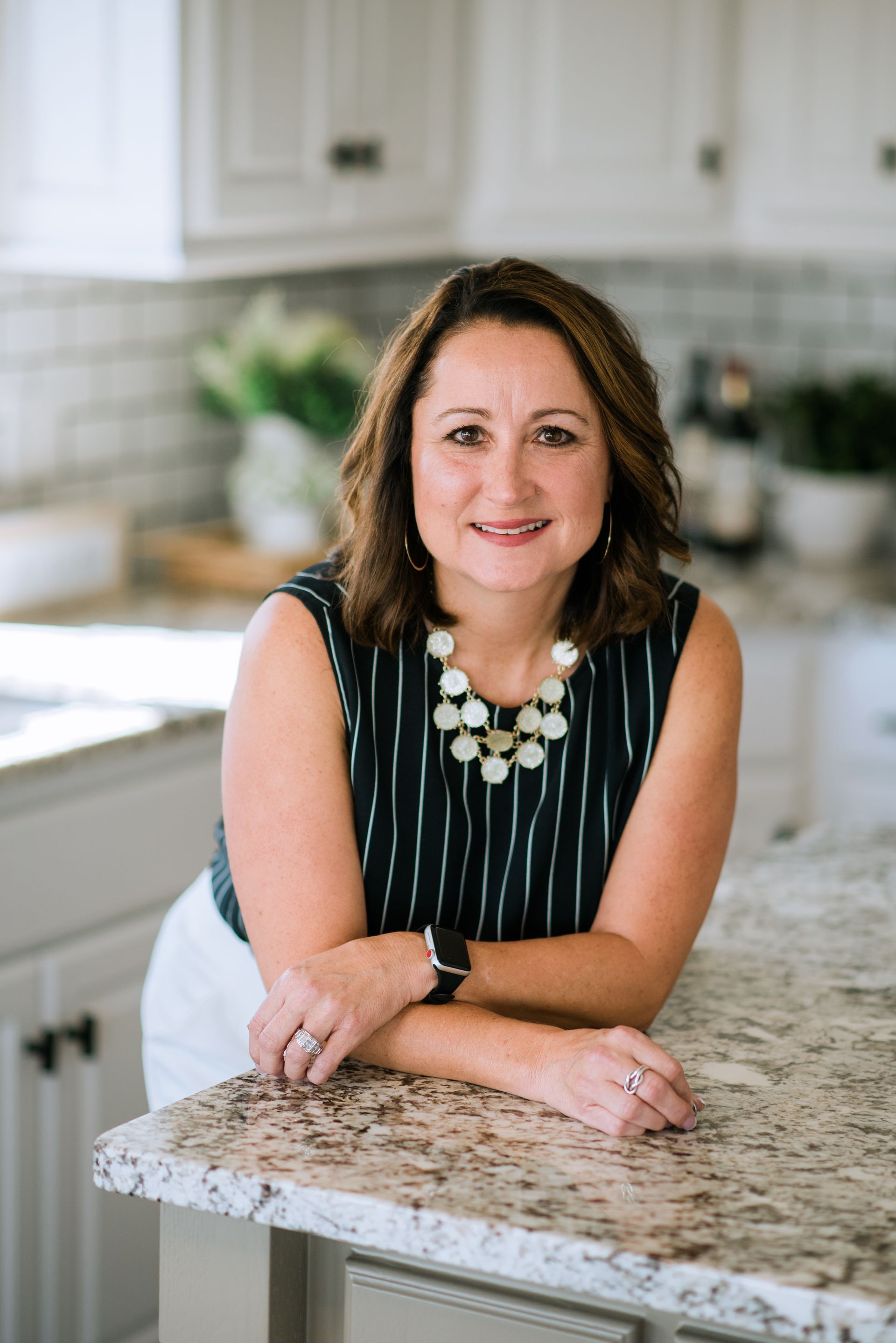 Woman leaning on a kitchen island in a bright white kitchen, wearing a striped top and statement necklace