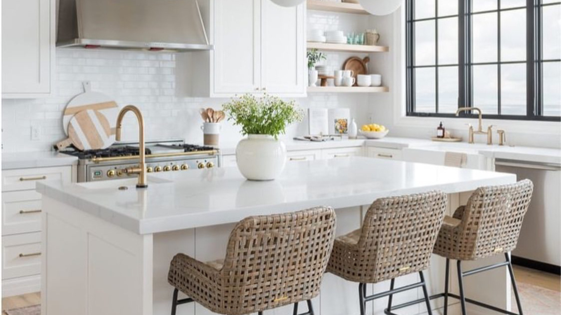 A kitchen with white cabinets , a large island , wicker chairs and a stove.