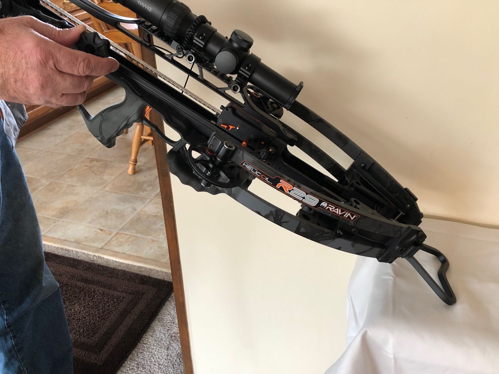 A person is holding a crossbow with a scope and Heart Drop Hanger attached to it