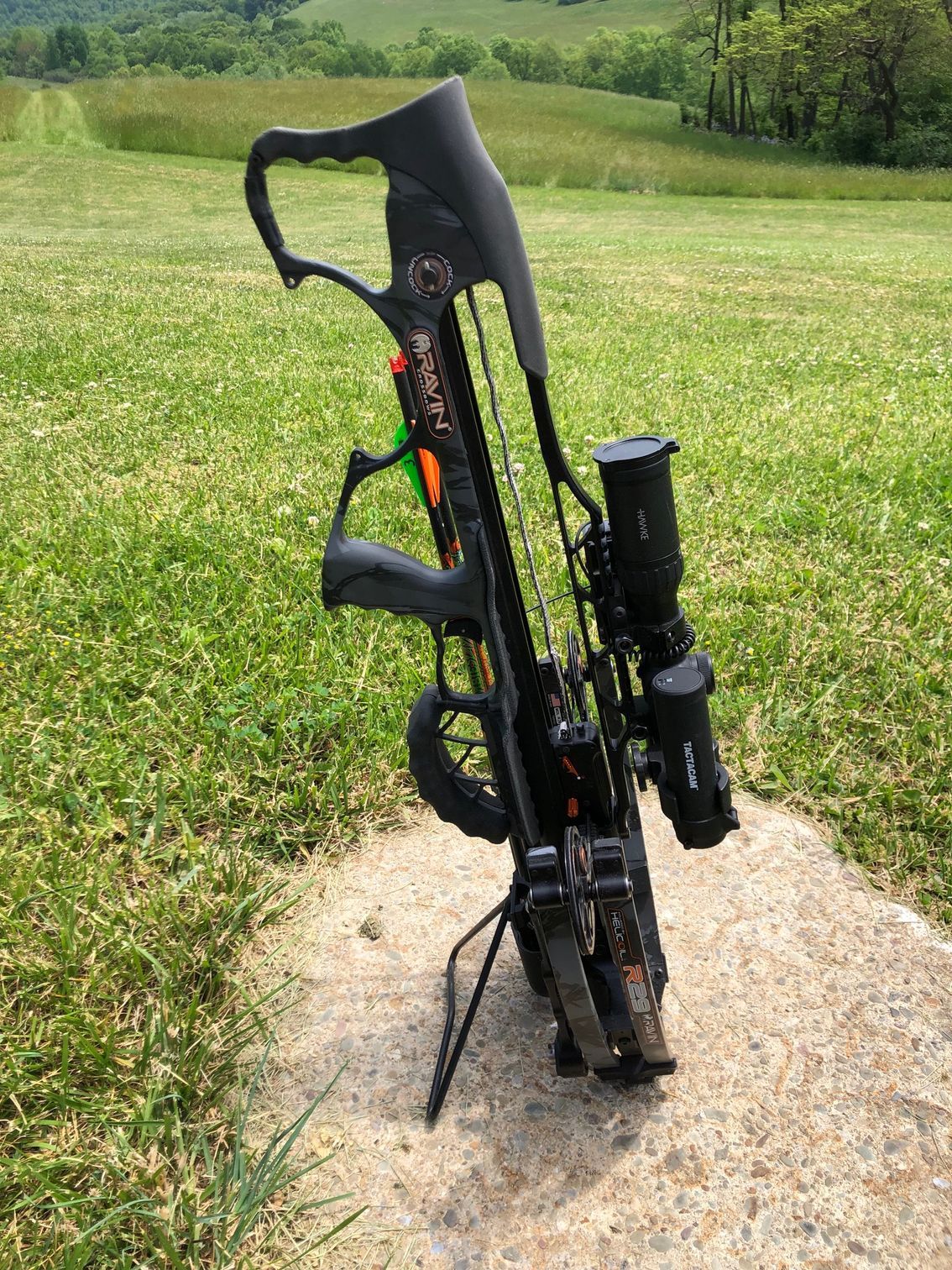A crossbow is sitting on top of a rock in a grassy field with the Backyard Broadheads kickstand enabled. 