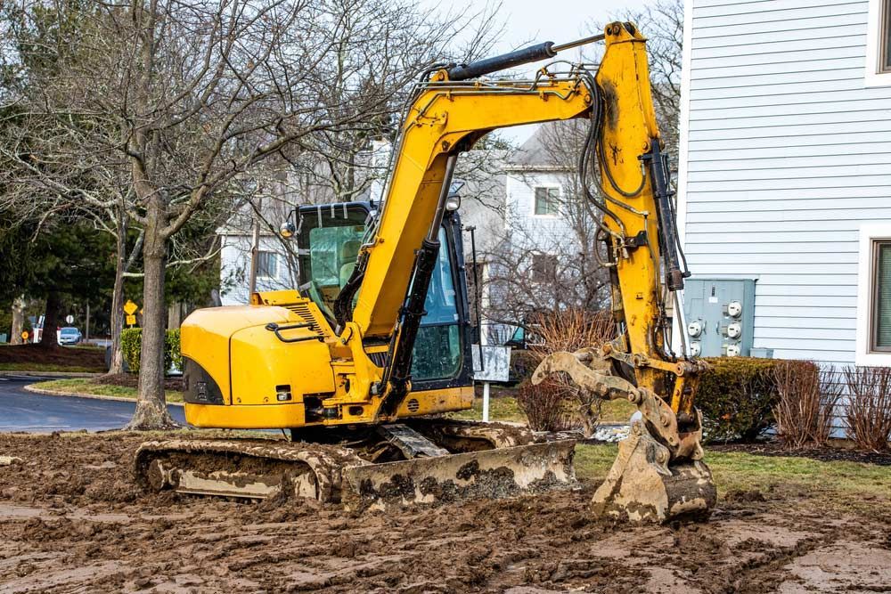 A Yellow Excavator is Digging a Hole in the Dirt in Front of a House — Tony Eldridge Property Services in Forster, NSW