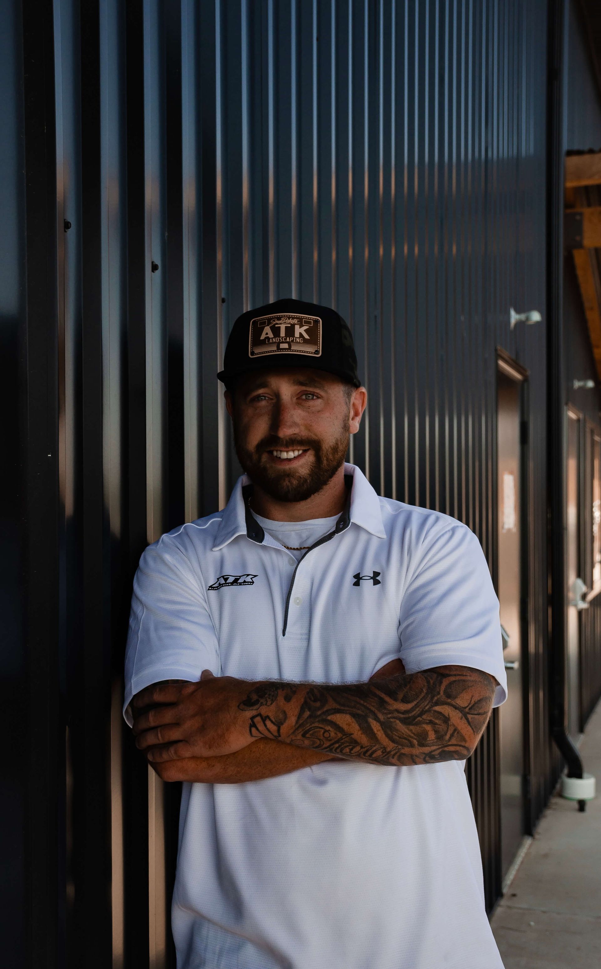 A man with a beard and tattoos is standing in front of a building with his arms crossed.