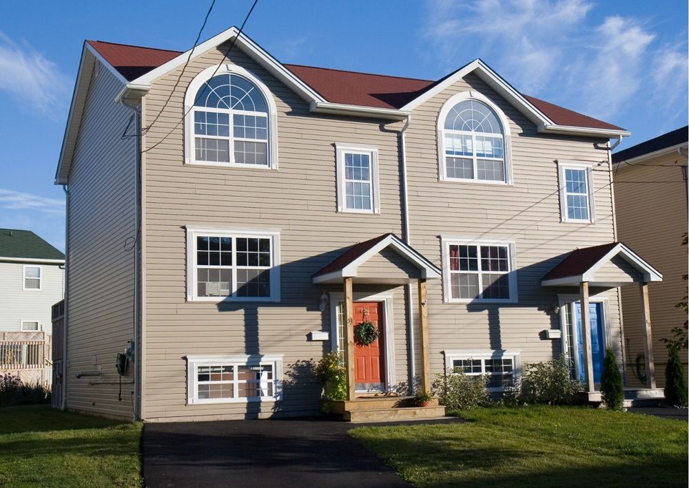 Tan vinyl sided semi-detached home with arched windows, burgundy roof, covered entry porches, and green lawn under clear sky.