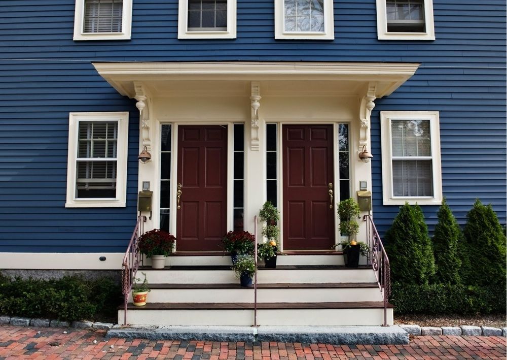 Navy blue colonial style duplex with white trim, twin brown doors, covered porch, and symmetrical windows with brick walkway.