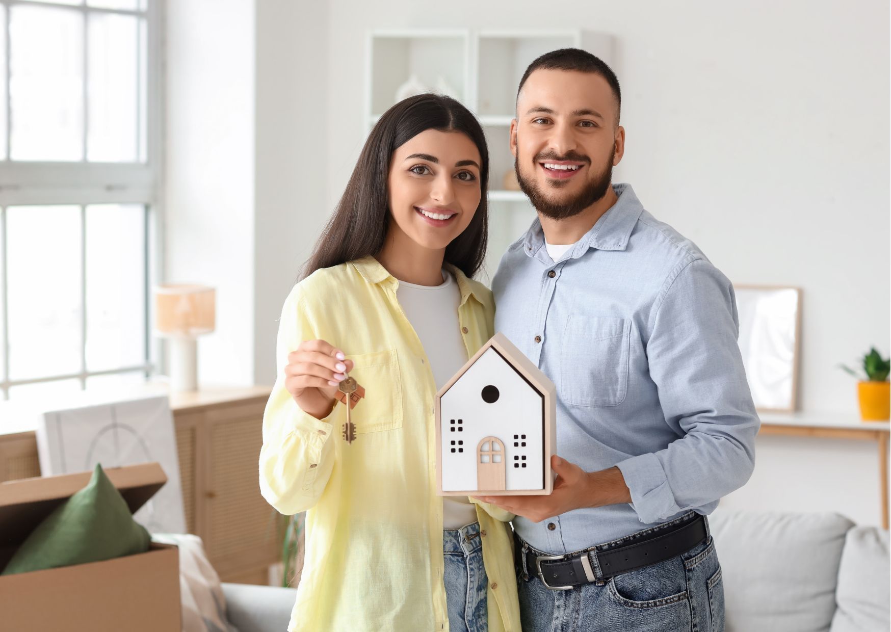 Smiling couple holding house keys and miniature house model in their new bright living space.