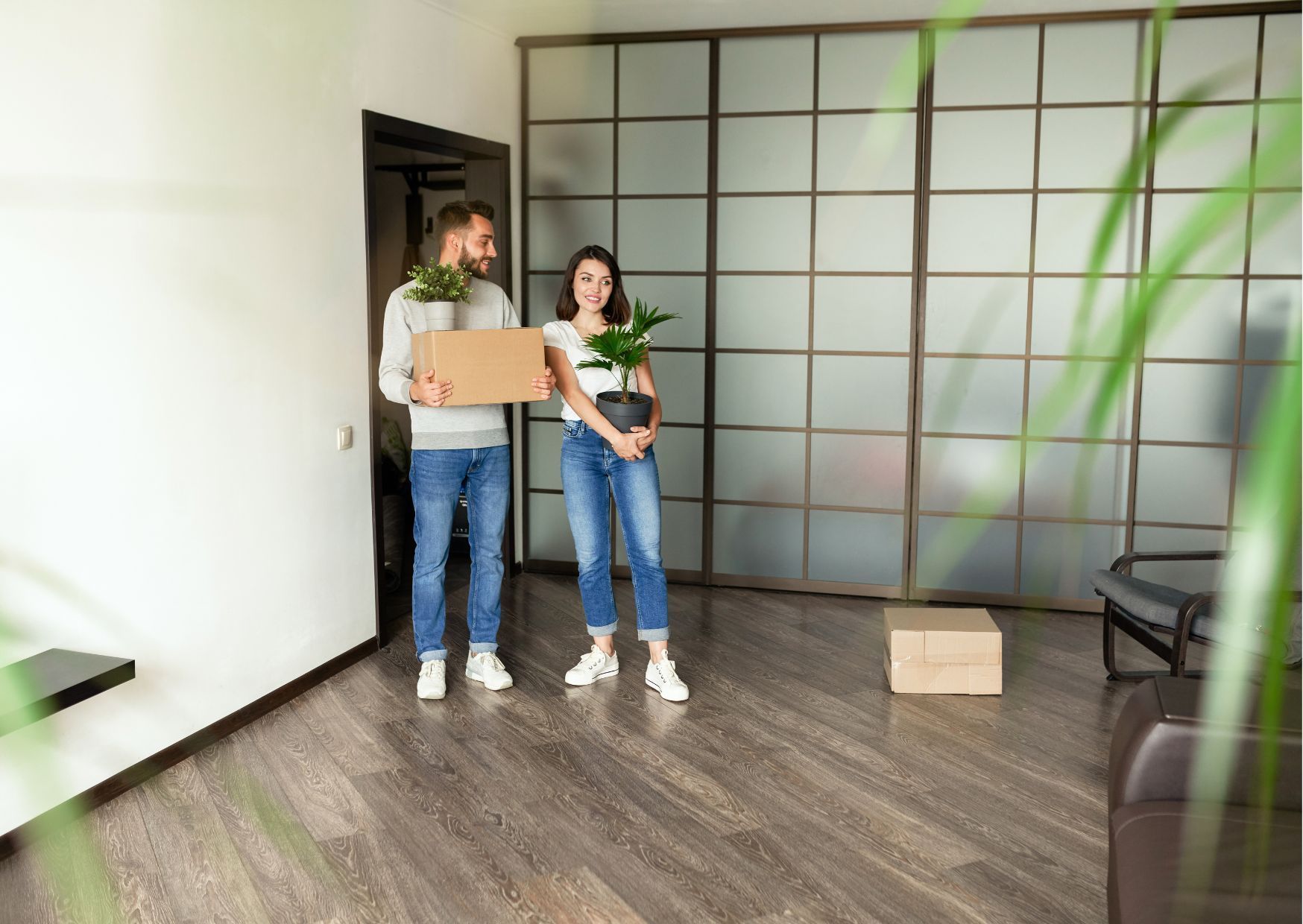 Young couple carrying moving boxes into empty apartment with modern glass partition and wooden floors.