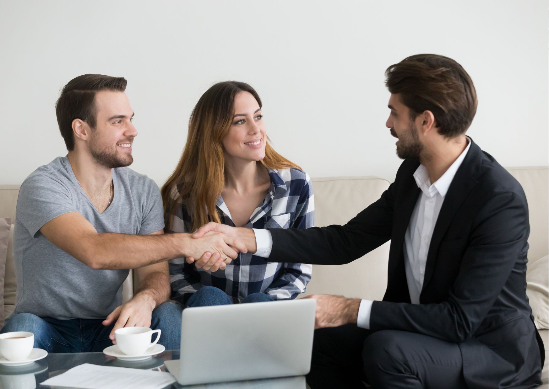 Real estate agent shaking hands with couple during property consultation meeting with laptop and coffee.