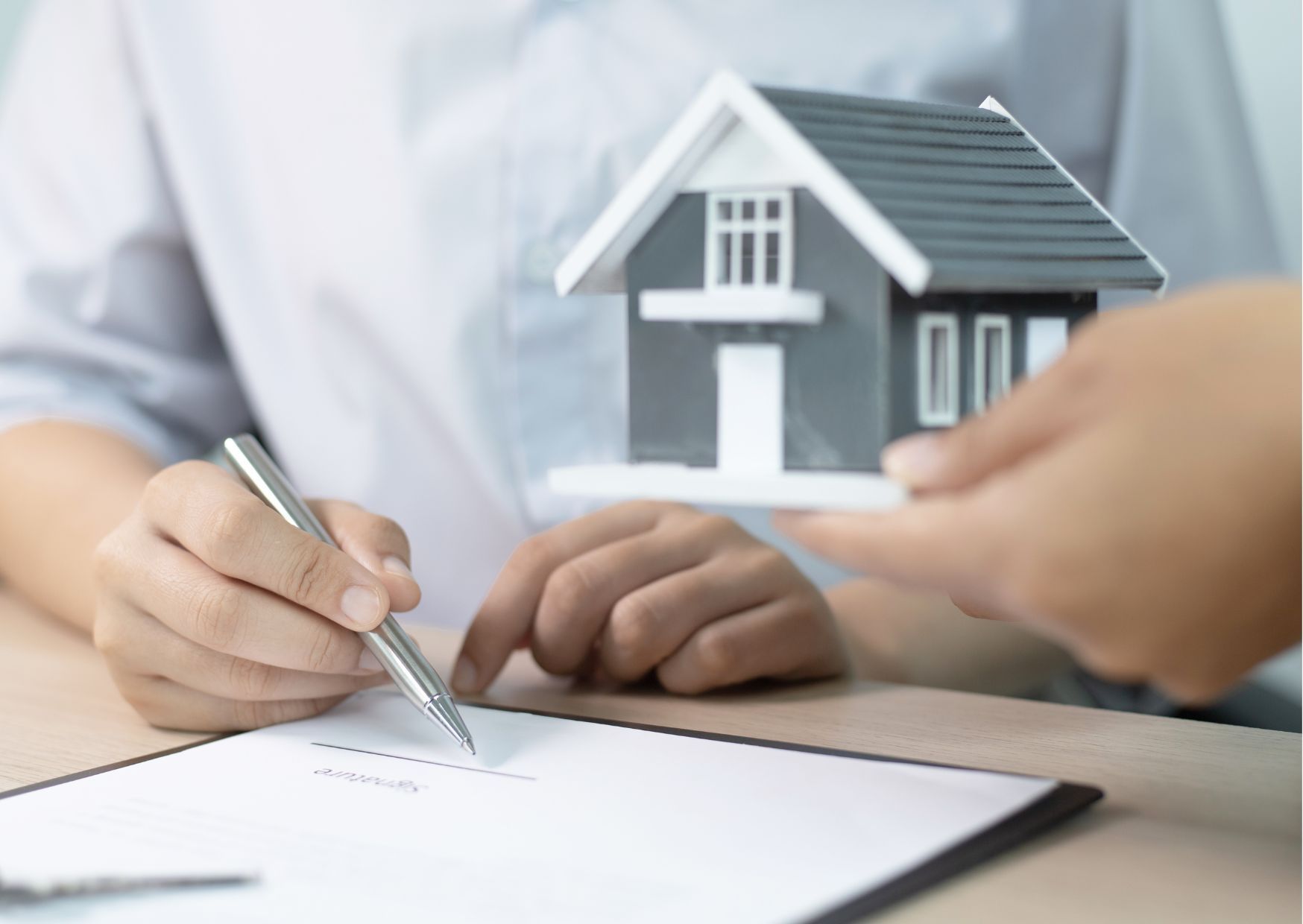 Hands signing rental contract document with house model and pen on desk during lease agreement.