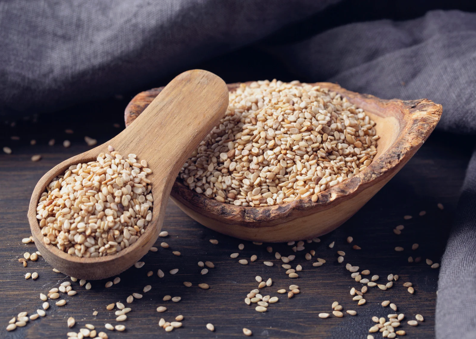 Wooden bowl and spoon overflowing with beige sesame seeds on a dark wood surface.