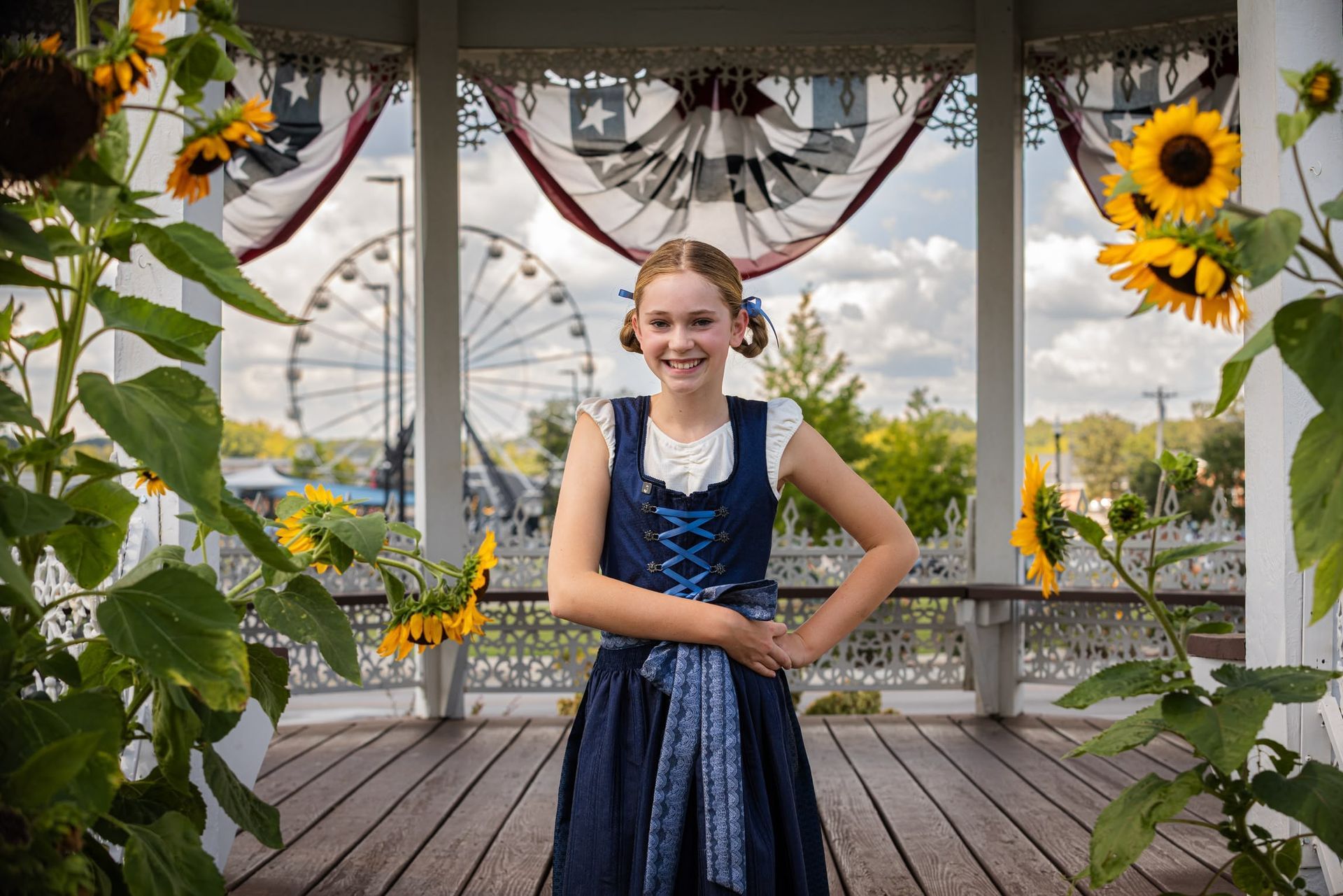 Smiling person in a blue dress standing on a wooden gazebo decorated with sunflowers and bunting.