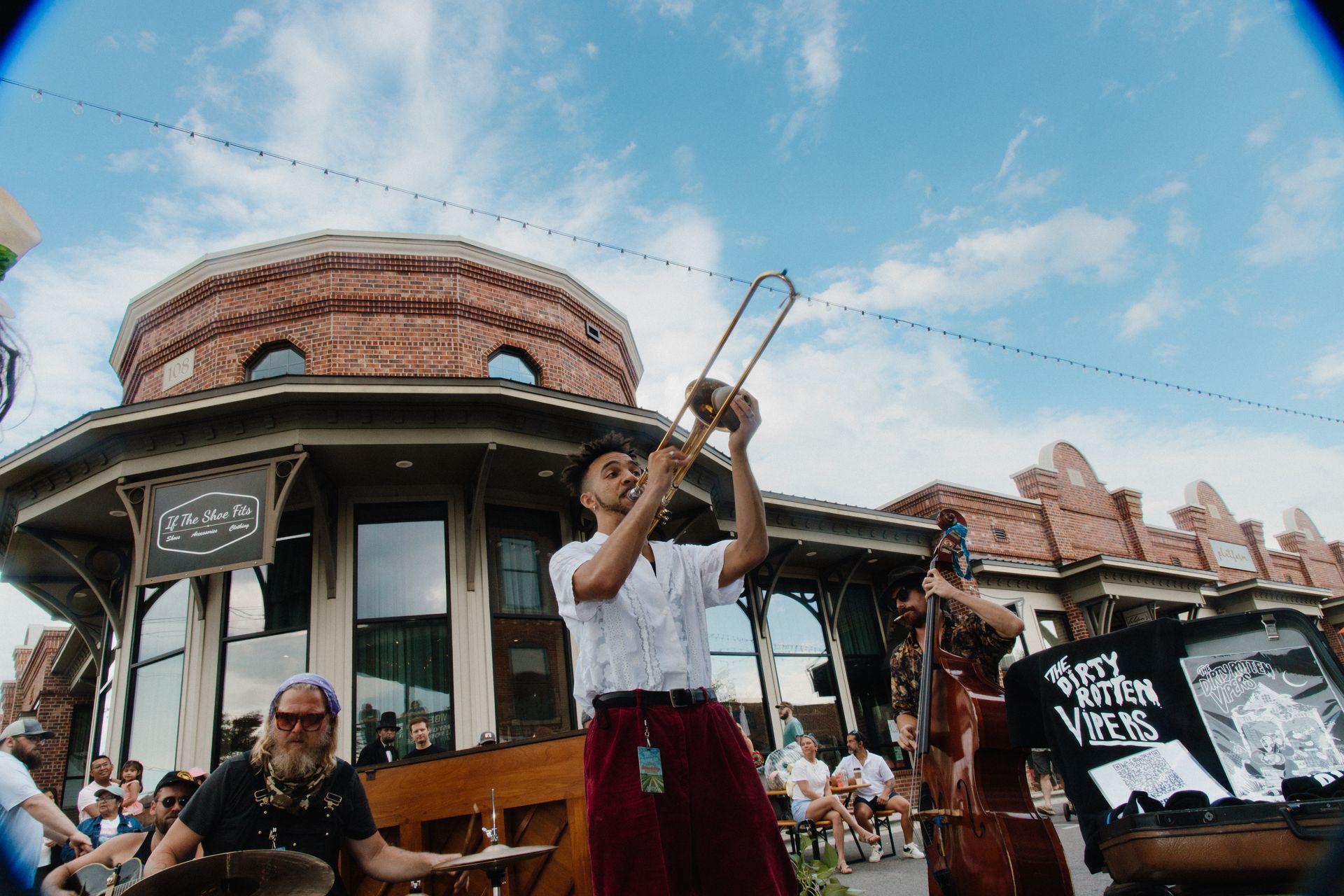 Musicians performing outdoors on a street, with a trombone player in front of a brick building under a blue sky.