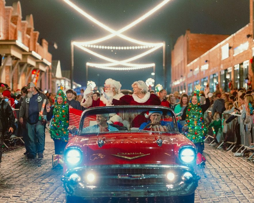 Santa in a red convertible amid a festive holiday street parade with crowd and string lights