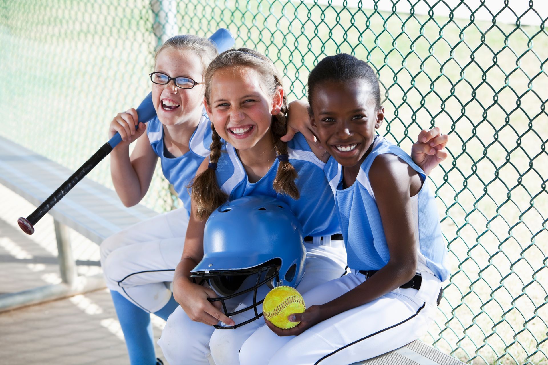 Three young girls are sitting next to each other in a dugout holding baseballs and bats.