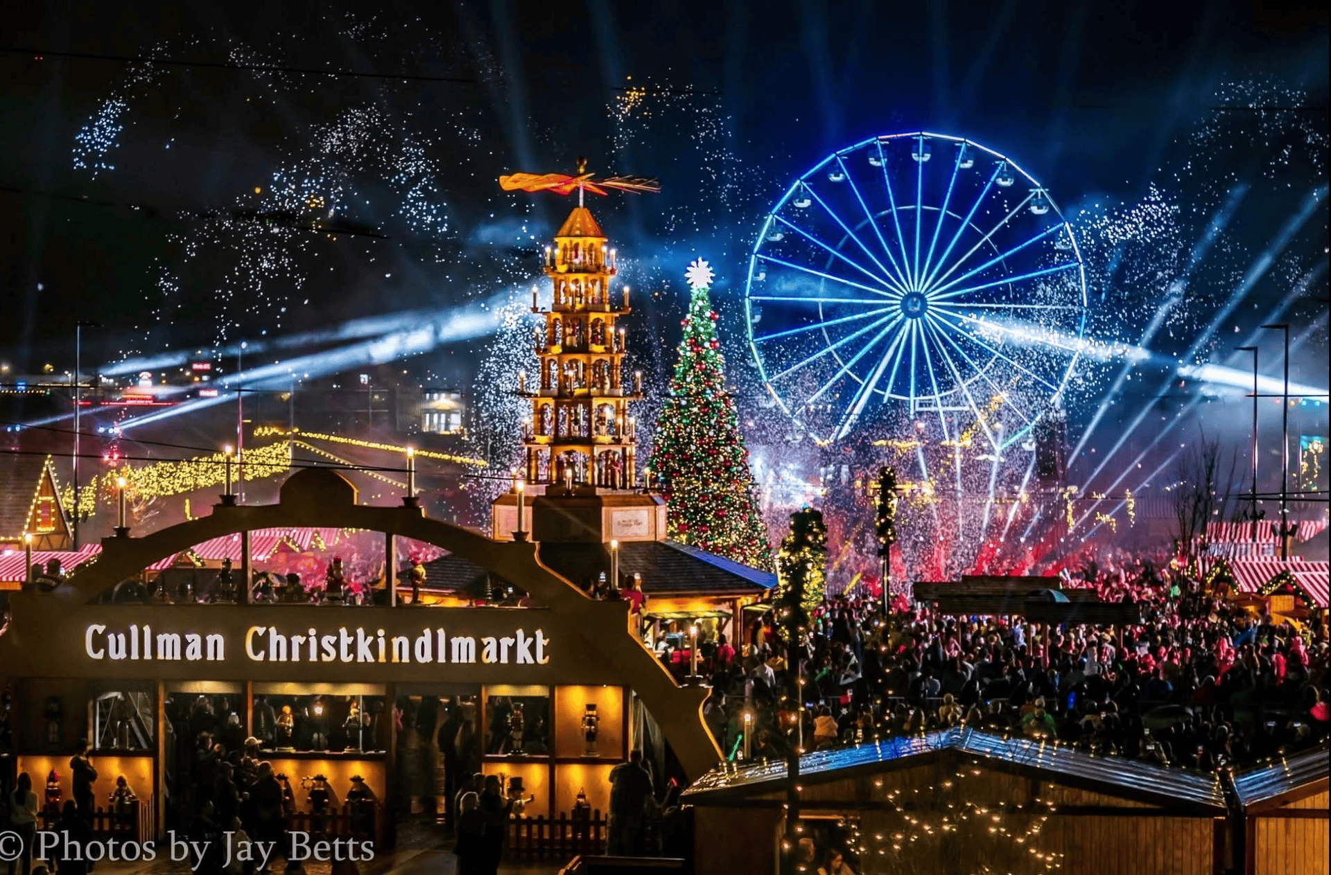 Festive Christmas market at night with lit tree, Ferris wheel, fireworks, and crowds