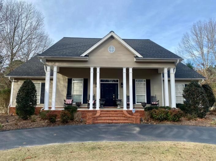 Front porch of a beige house with white columns, dark roof, and brick steps, framed by shrubs and bare trees