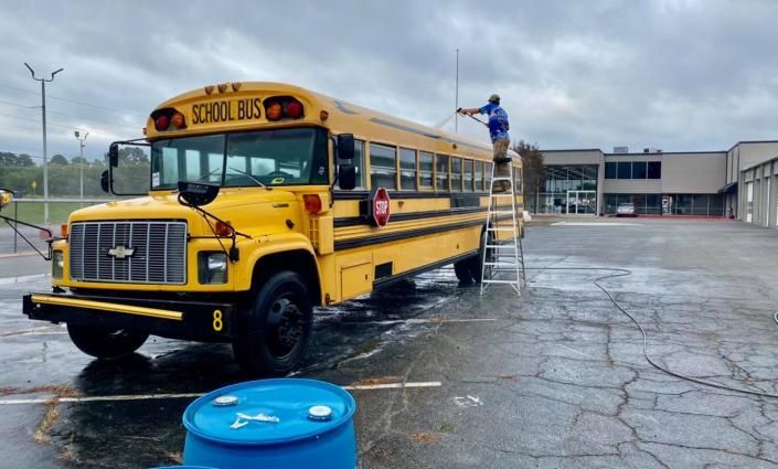 Yellow school bus being washed in a wet parking lot, with a person on a ladder cleaning the roof.