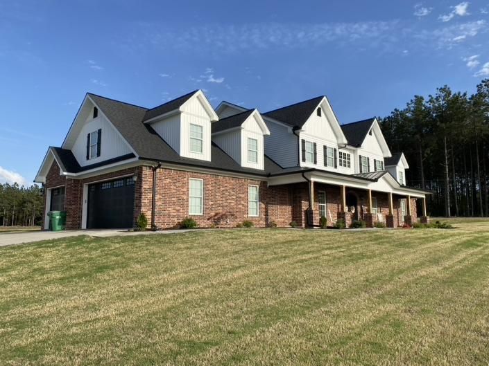 Large brick house with gray roof and dormer windows on a grassy hill under a blue sky