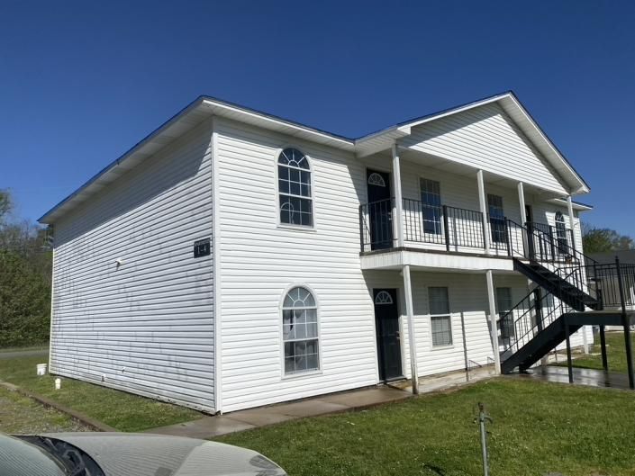 White two-story apartment building with black balconies and exterior stairs on a grassy hillside