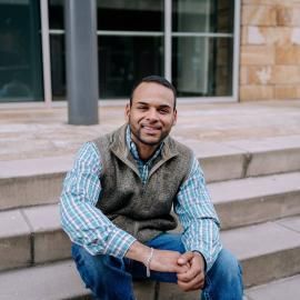 Man sitting on steps outside a building, smiling with hands clasped, wearing a plaid shirt and vest.