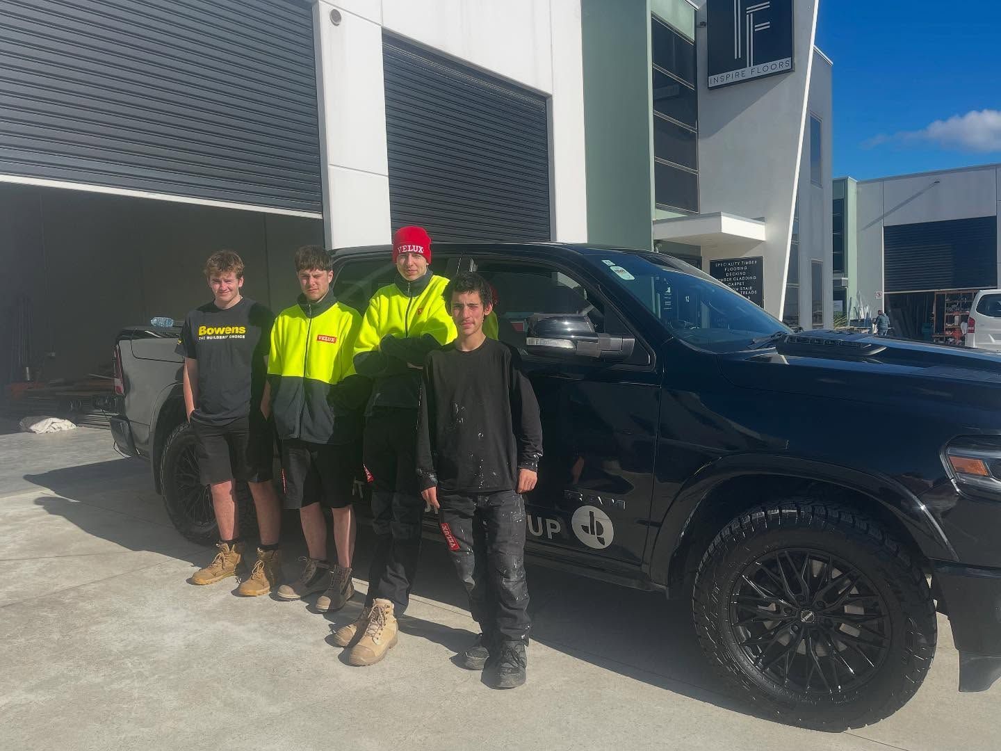 Four people standing beside a black pickup truck outside a building in bright sunlight