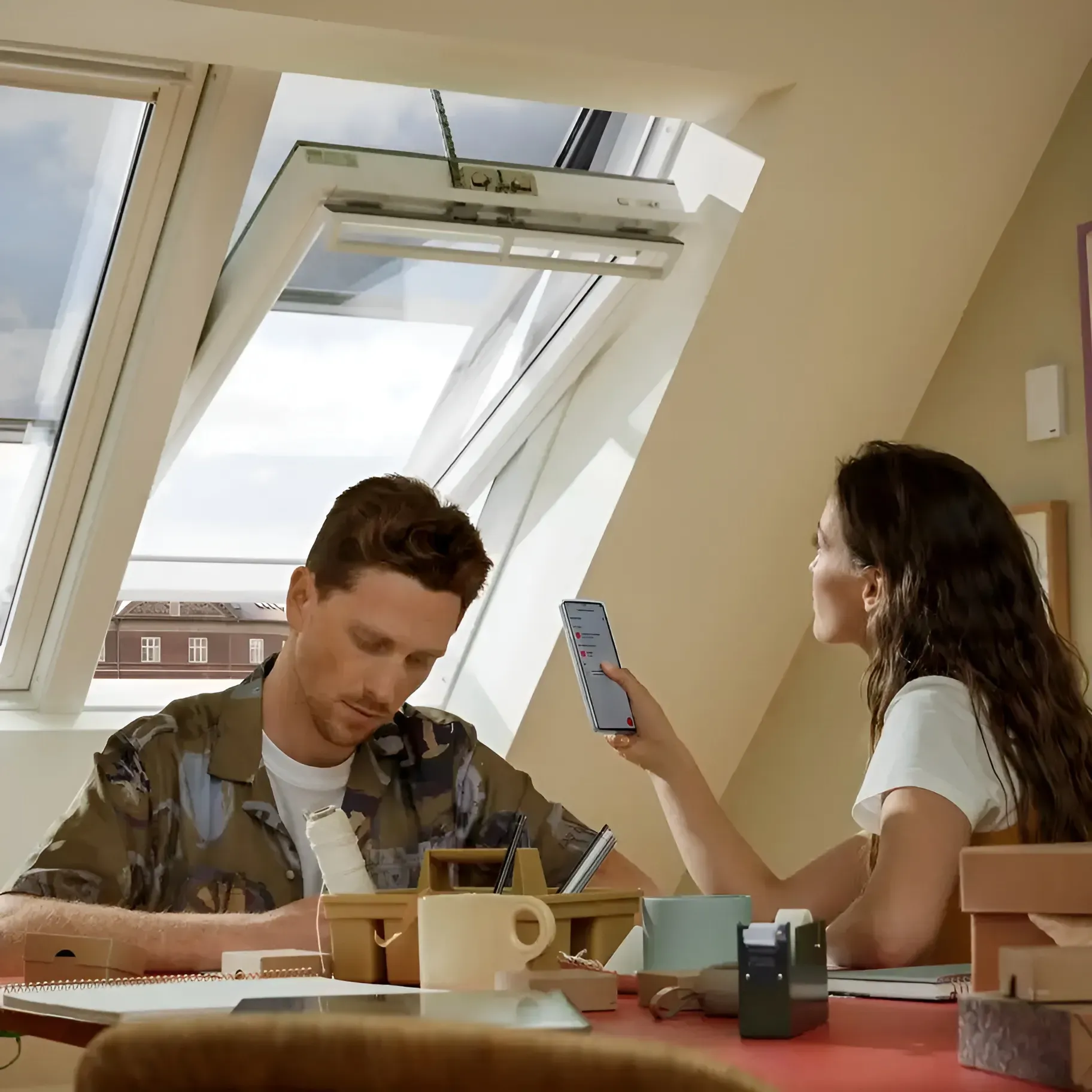 Two people sit at a table in a bright room, one reading a phone and the other writing.
