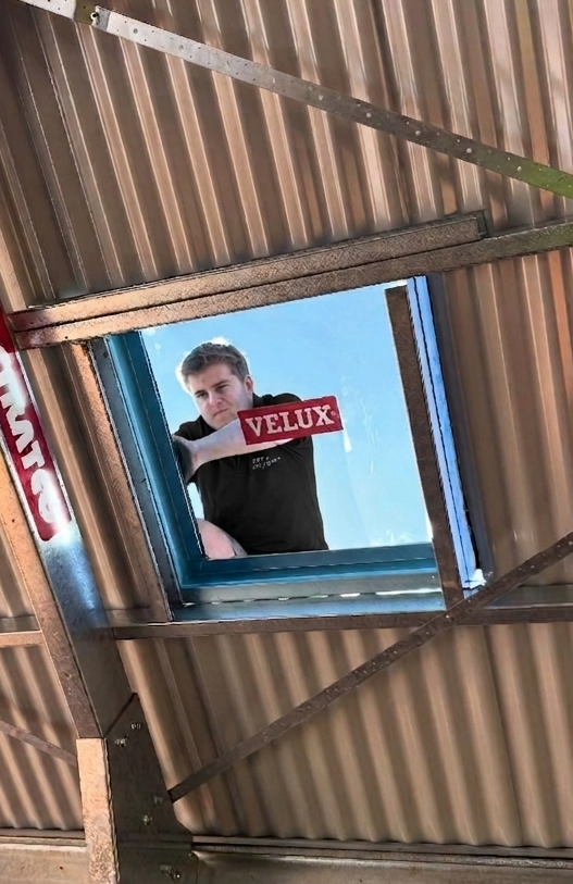 Man standing outside a VELUX roof window in a corrugated metal roof, with blue sky behind him