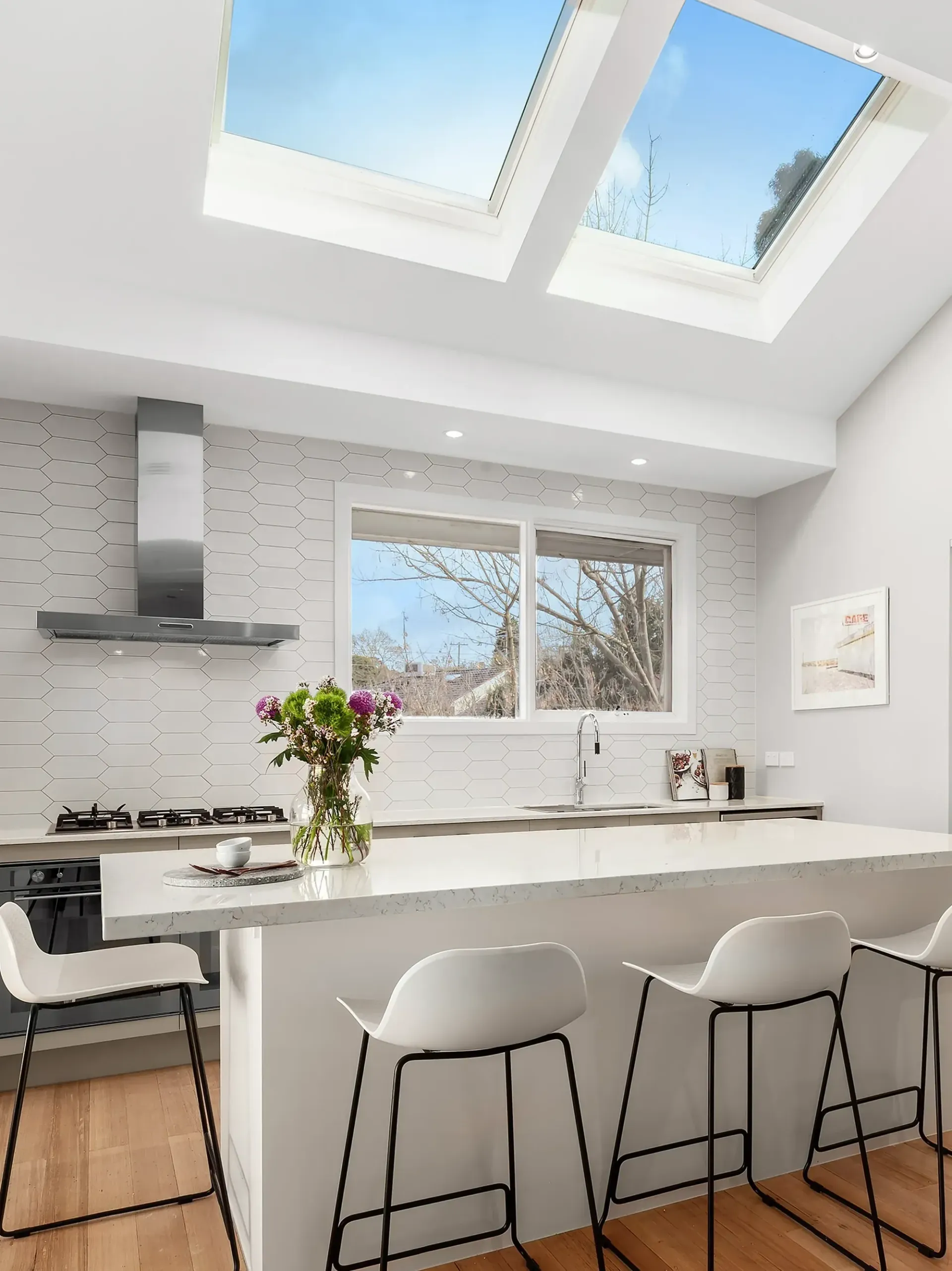 Bright modern kitchen with skylights, white island, bar stools, and a vase of flowers.