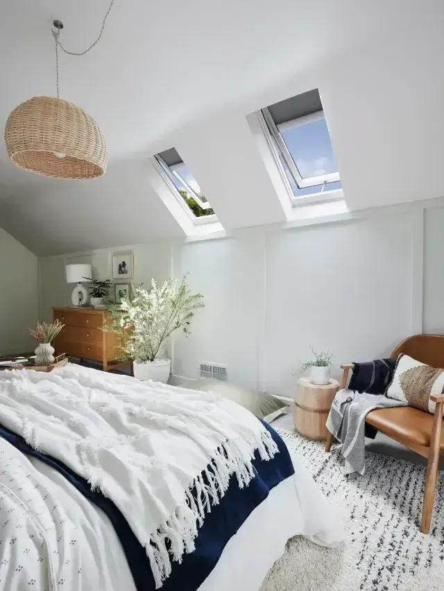 Bright attic bedroom with sloped skylight windows, white bedding, woven pendant lamp, and wooden chair.