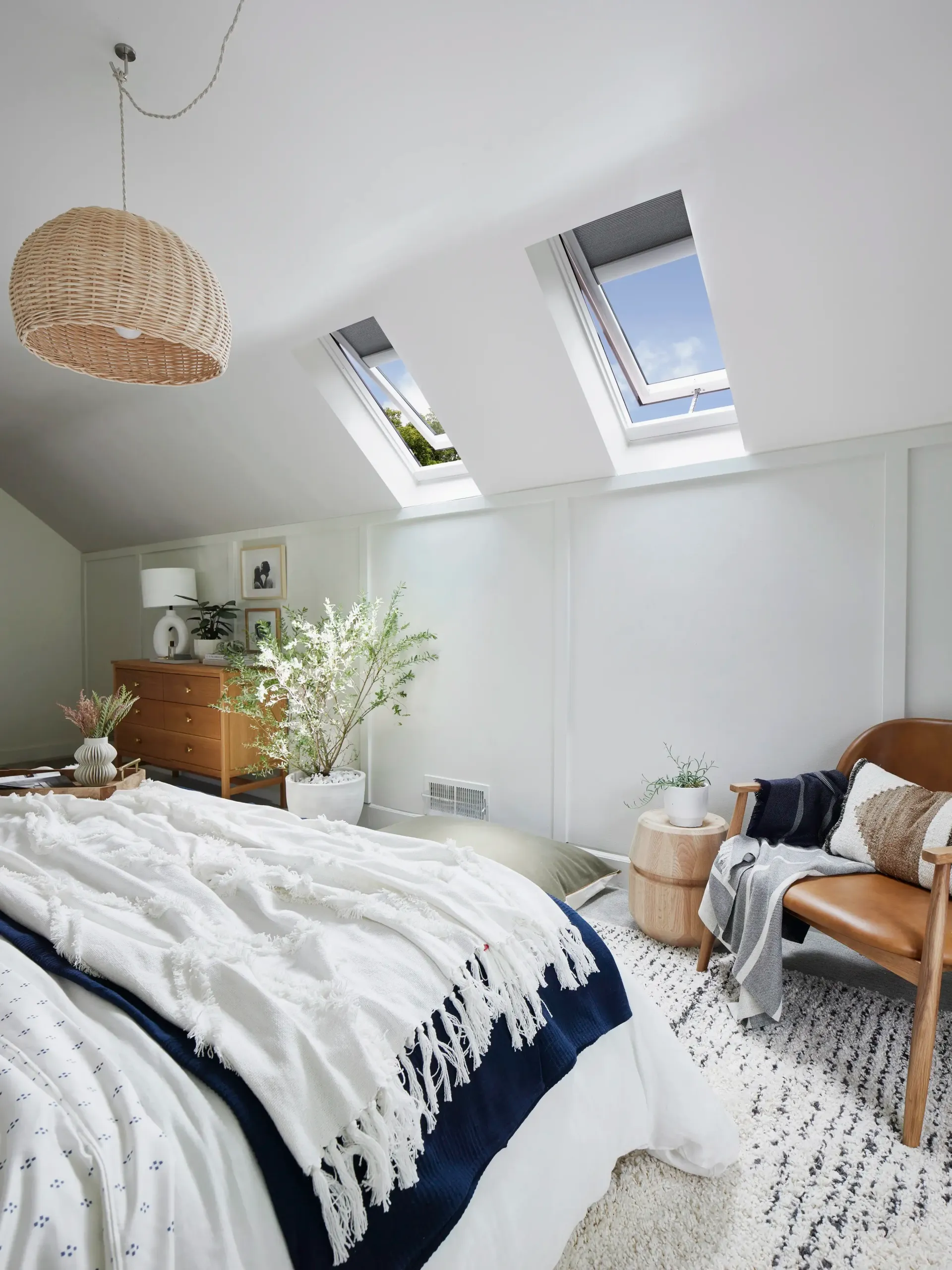 Bright attic bedroom with sloped skylight windows, white bedding, woven pendant lamp, and wooden chair.