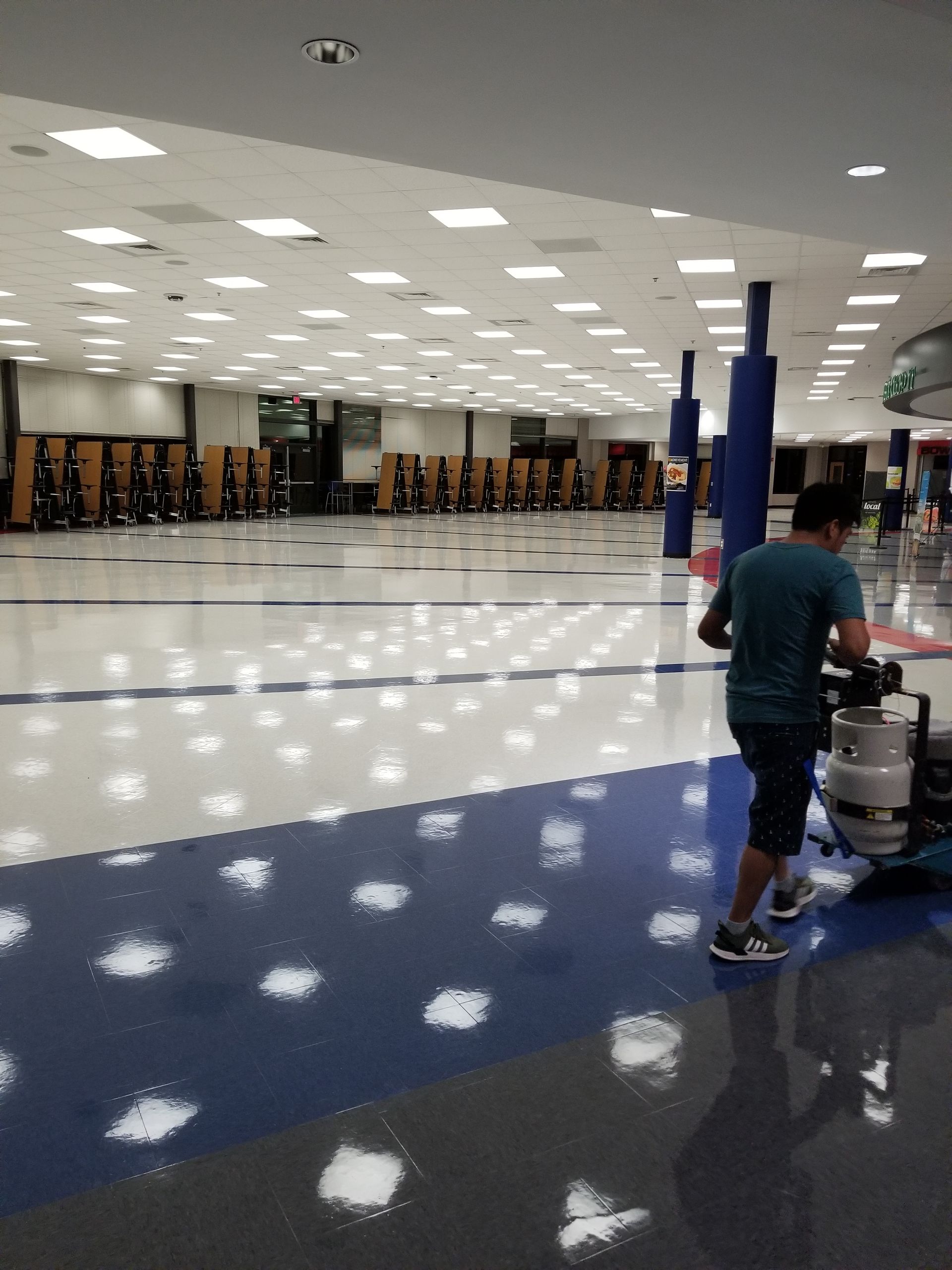 Bright indoor hall with polished floor, overhead lights, and one person pushing a cart near the right side.