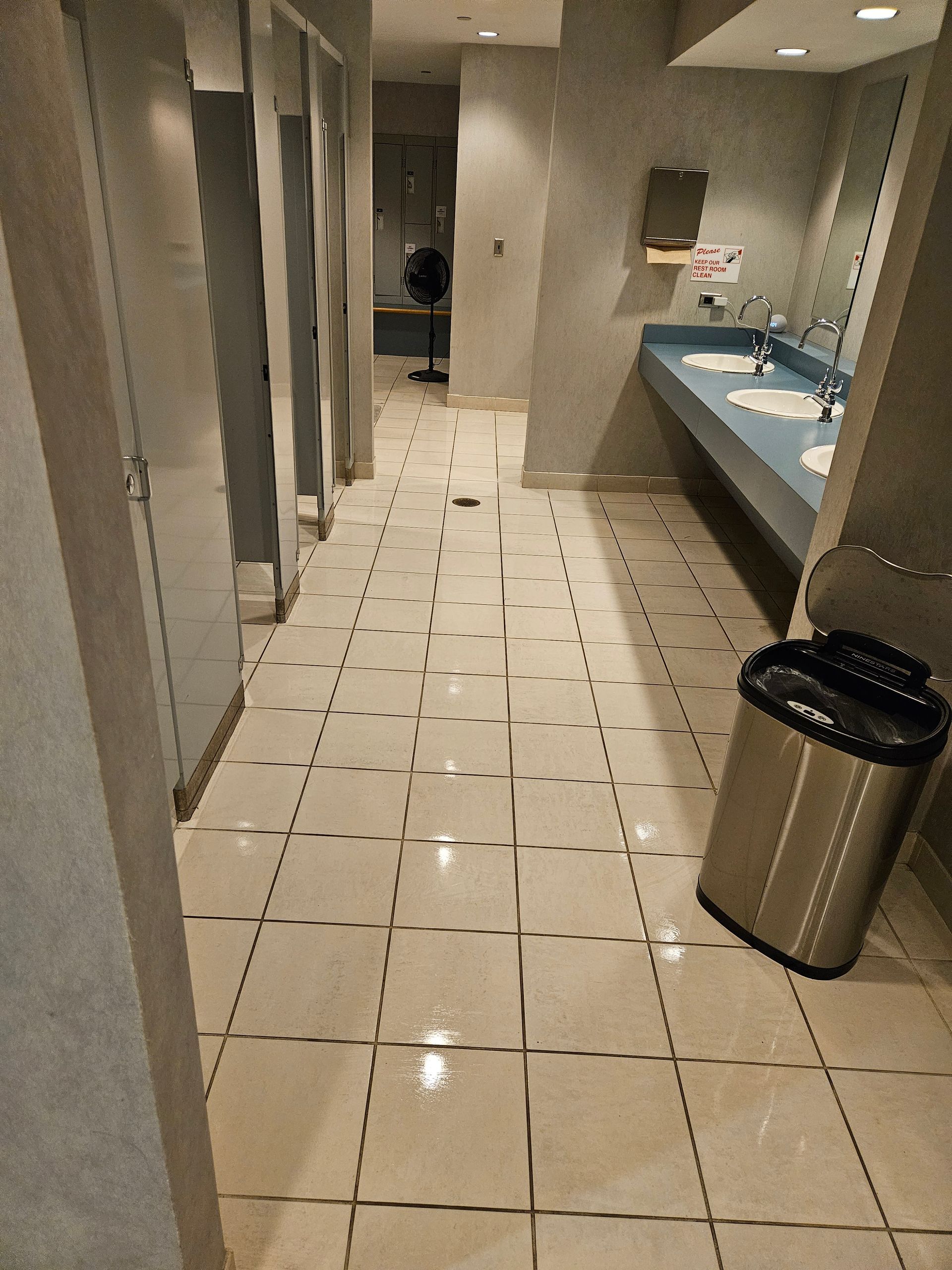 Public restroom hallway with tiled floor, stalls on the left, sinks on the right, and a trash can.