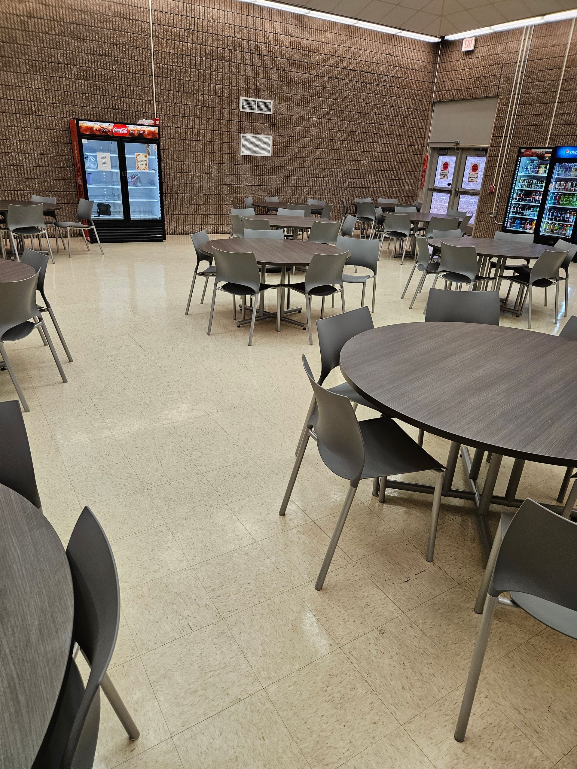 Empty cafeteria with round tables and chairs under a textured brown wall, bright vending machines in the back.
