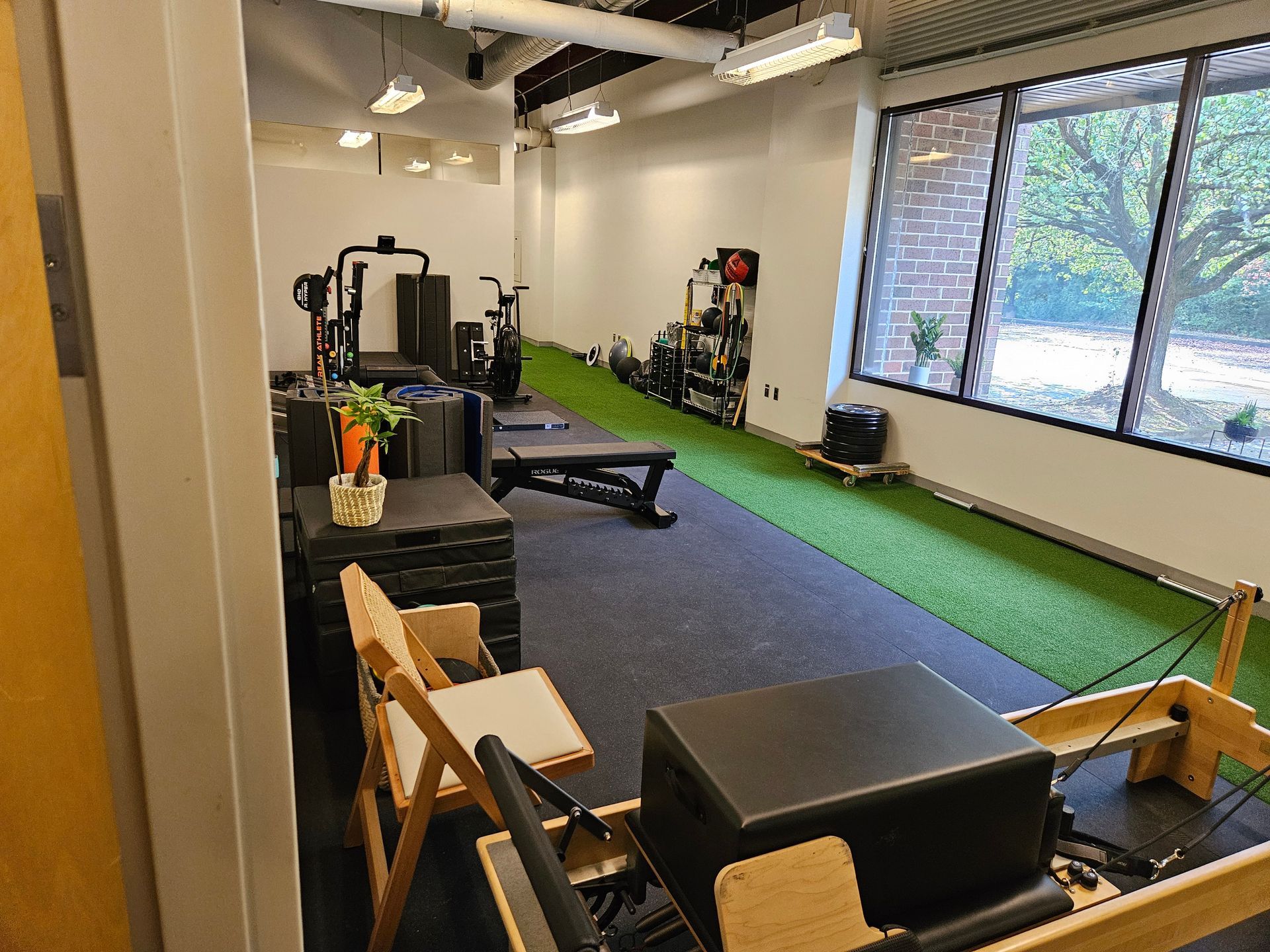 Empty gym studio with exercise machines, green turf, large windows, and folding chairs.